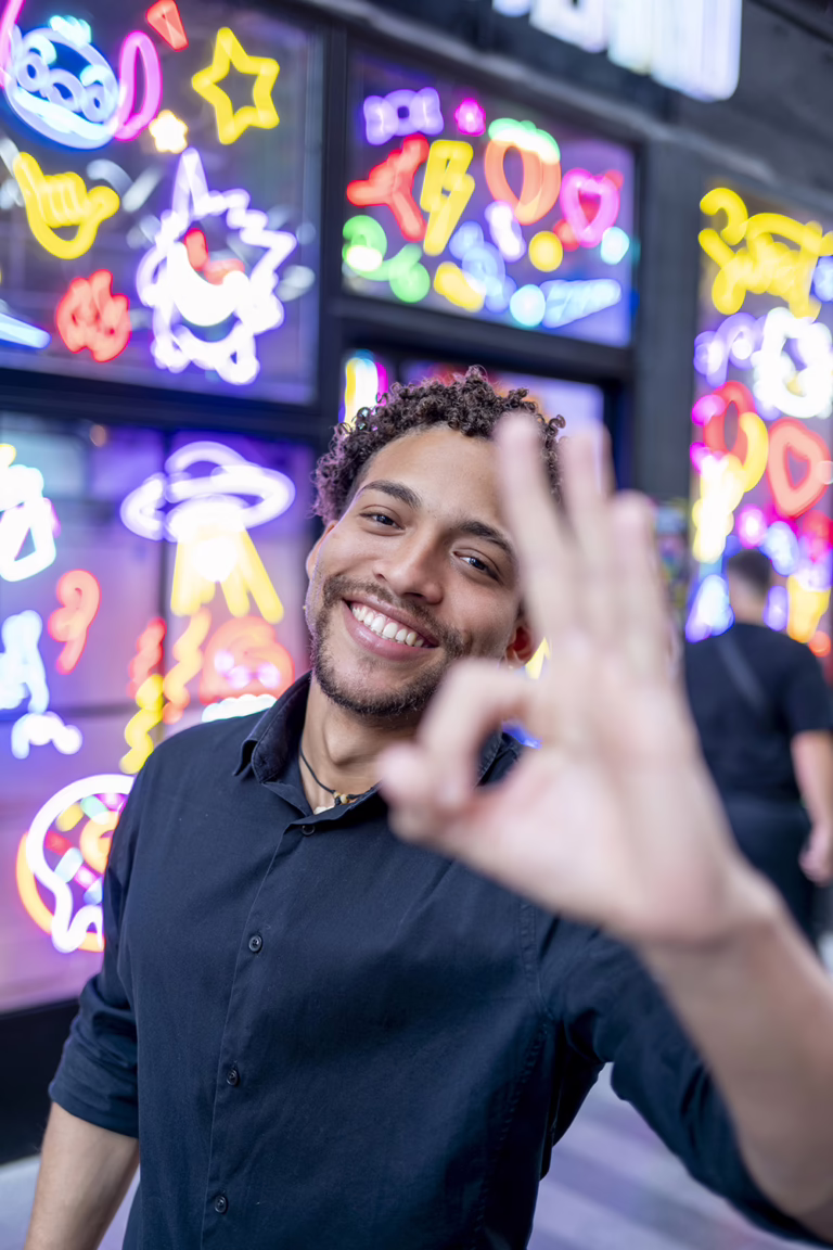Smiling man in a dark shirt gives an OK sign toward the camera with bright neon signs in the background.