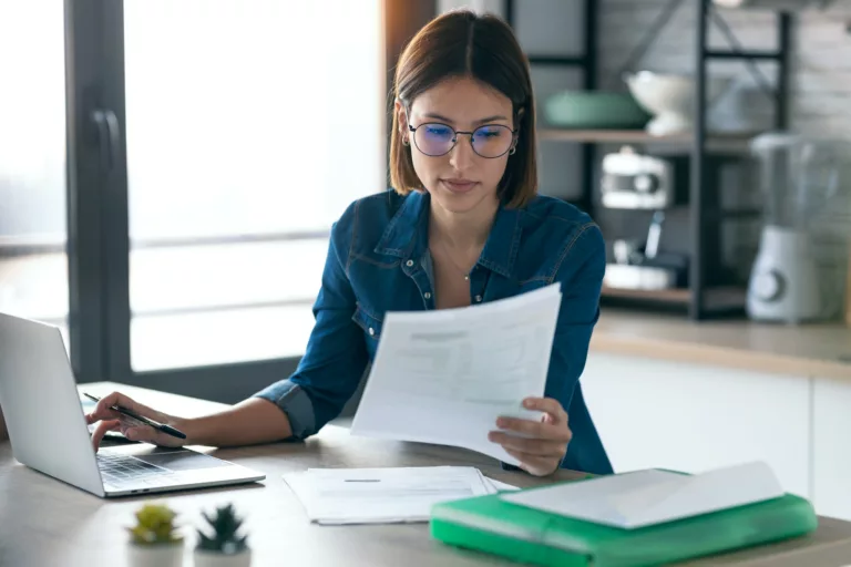 young woman working with computer while consulting some invoices and documents in the kitchen. Image for AP Automation pages of the Document Solutions section of the MCC website