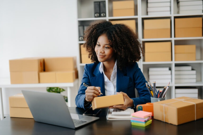 Woman in a blue blazer packs a small box at a desk with a laptop and shelves full of boxes in the background.