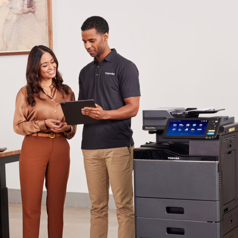 Two coworkers stand by a Toshiba multifunction printer, looking at a tablet and discussing.