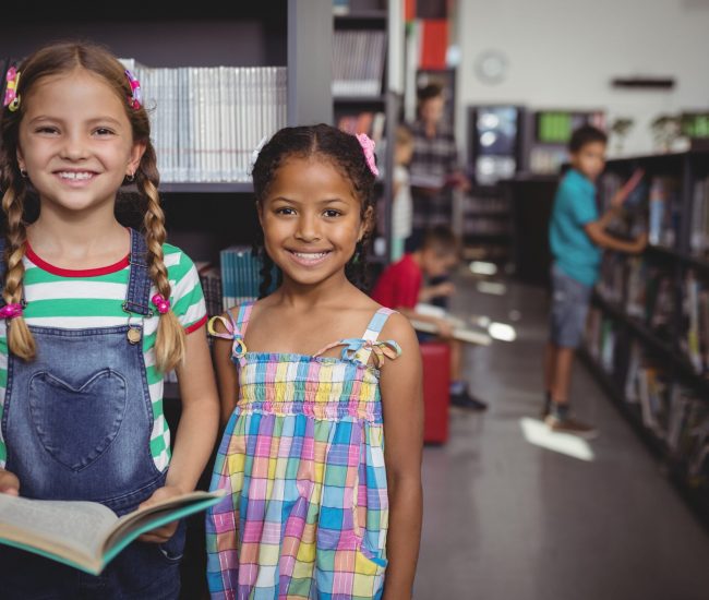 Two smiling young girls stand in a library by a bookshelf; one holds an open book as they pose for the photo.