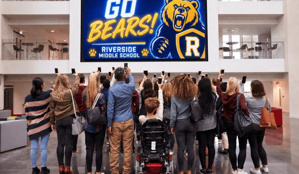 Group of students and a person in a wheelchair stand with raised phones, facing a large digital screen that reads 'Go Bears! Riverside Middle School' with a bear mascot in a school lobby.