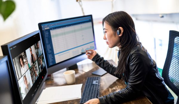 Support agent with headset at a desk, dual monitors showing video tiles and a data dashboard on the right.