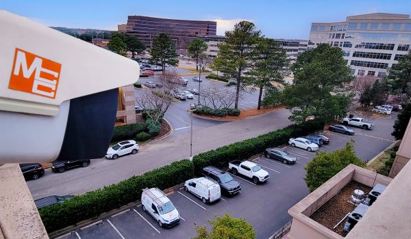 MCC Commercial CCTV Security Camera mounted to the top of a building overlooking a parking lot full of cars.