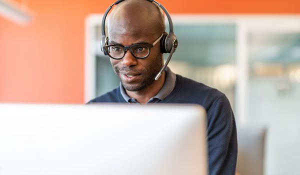 Man wearing over-ear headset and glasses, focused on a computer screen in an office with orange wall.