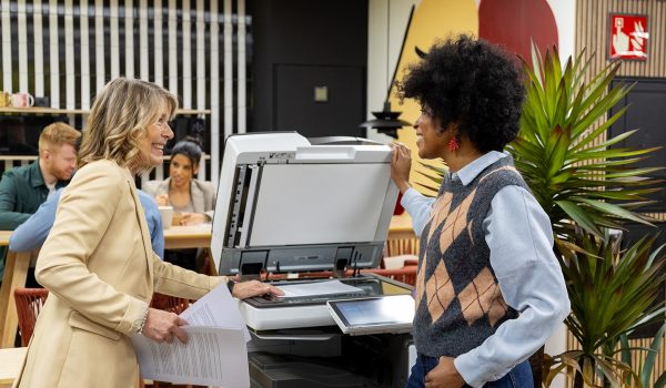 Two colleagues in a bright office near a photocopier, exchanging documents and chatting.