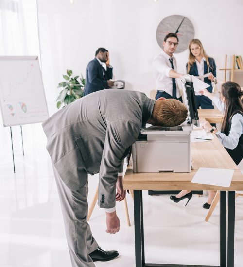 Man in a gray suit bends over a printer on a conference desk as coworkers debate documents in the background.