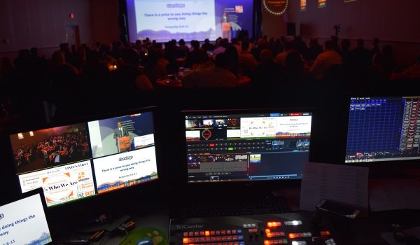 Image featuring computer screens in the foreground, displaying the NewTek Tricaster video production system in operation. In the blurred background, a large event is taking place, hinting at the real-world application of the advanced technology. This image showcases the seamless integration of high-tech video production into live events.