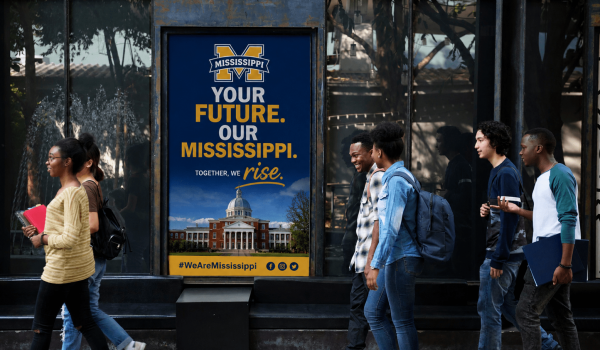 A group of diverse students walk past a large blue Mississippi poster that reads 'Your Future. Our Mississippi' with a campus building image at the bottom.