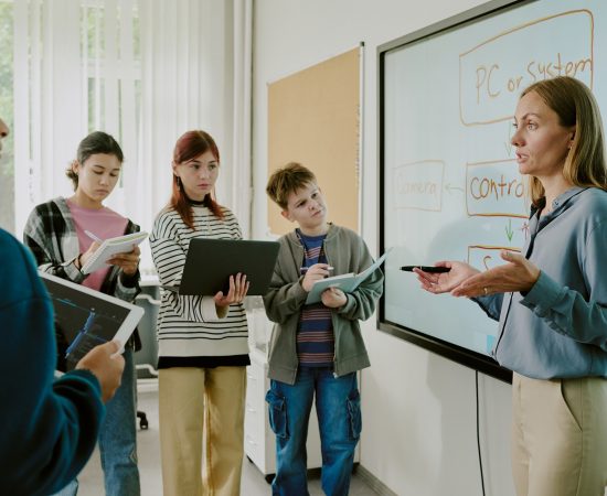Teacher explains a diagram on a large whiteboard as students listen, some holding notebooks.