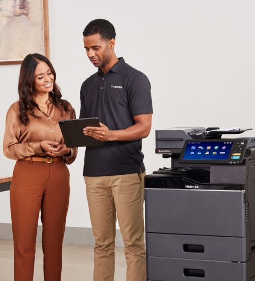 Two coworkers stand by a Toshiba multifunction printer, looking at a tablet and discussing.