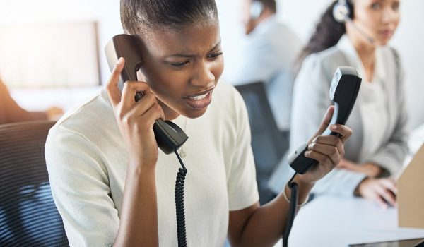 Customer service agent in a call center holding a phone to her ear while inspecting a second handset at her desk.