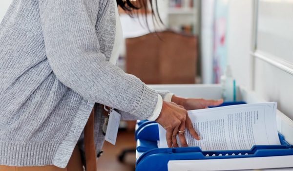 Person in a gray sweater placing papers into a blue document tray on a desk in an office setting.