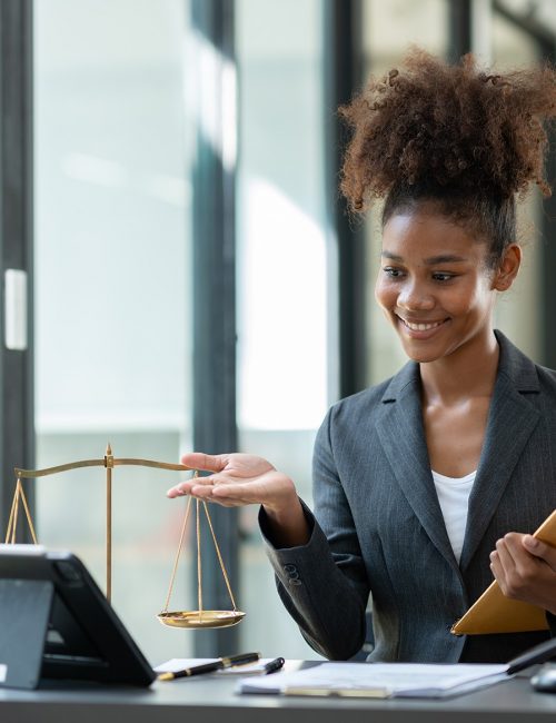 Smiling businesswoman in a blazer holds scales of justice at her desk, with documents and a gavel nearby.